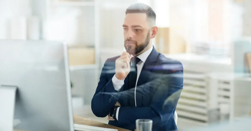 A businessman in a suit pondering deeply while looking at a computer screen, perhaps strategizing or problem-solving. Budget Analyst Jobs