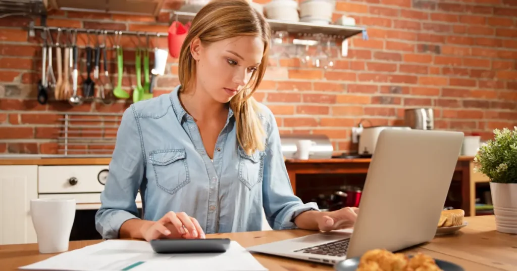 A woman calculating finances at home, using a calculator and laptop in the kitchen. Budgeting Behavior