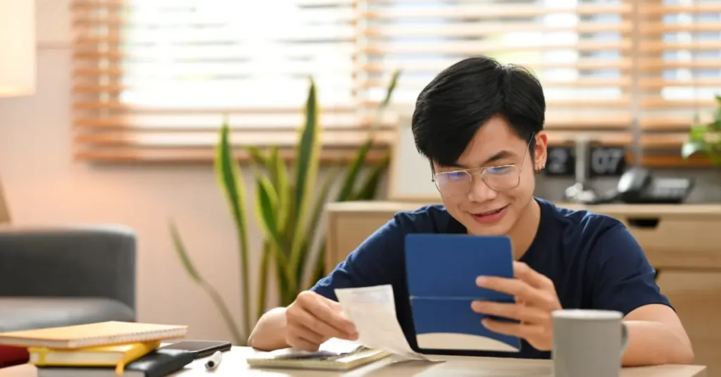 A teenager at a desk using a calculator and holding a bill, suggesting personal finance management. He is tracking his expenses in his budget worksheet for teenagers. 