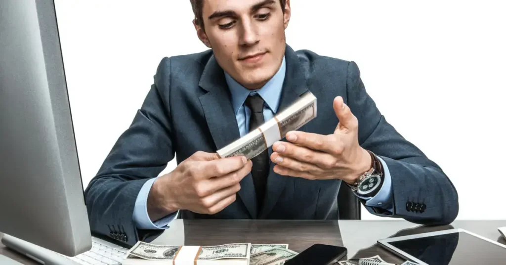 A man in a suit inspecting a bundle of cash at his desk with electronic devices around. It is the salary for the finance director job.