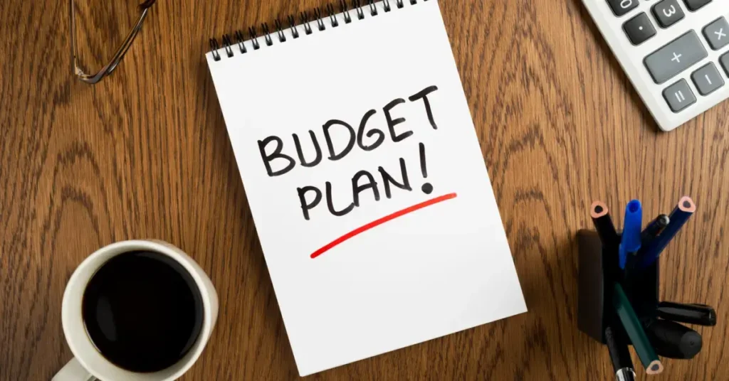 A notepad with "BUDGET PLAN!" written on it, a cup of coffee, glasses, and office supplies on a desk. Start setting up your budget worksheet for teenagers.