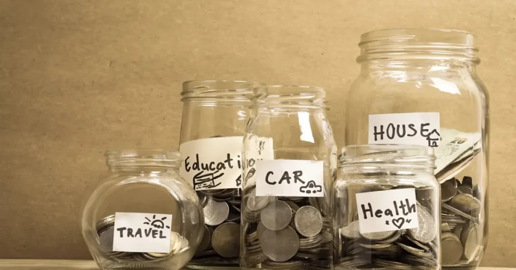 Glass jars labeled for different savings goals like "Travel," "Car," "House," and "Health," filled with coins. Try starting your s.m.a.r.t. goals after our budget workshop.