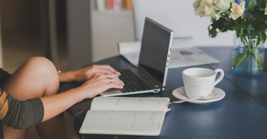 A person working on a laptop with a notebook and a cup of coffee on the table, suggesting remote work or study. The preson might started their remote finance jobs