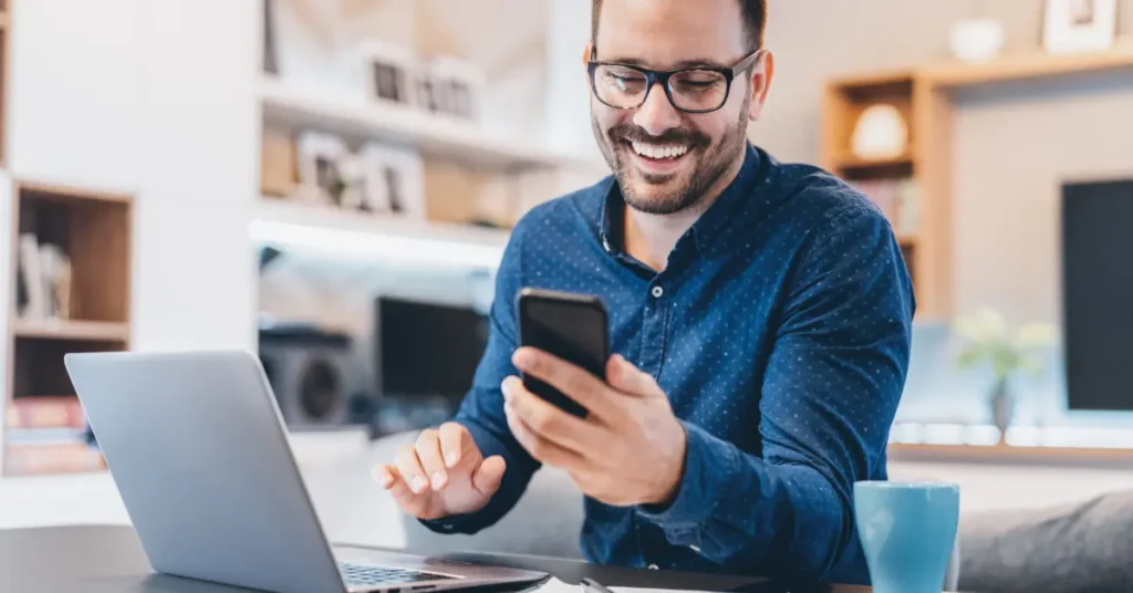 The same man laughing and looking at his smartphone while working on a laptop, possibly enjoying a break or good news. He is happy with his remote finance job.