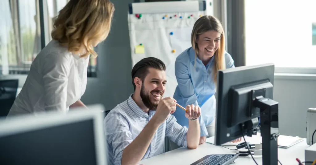 Three colleagues, two women and a man, smiling and looking at a computer screen, implying collaboration or a positive work discussion. Something you will not see while doing a remote finance job.