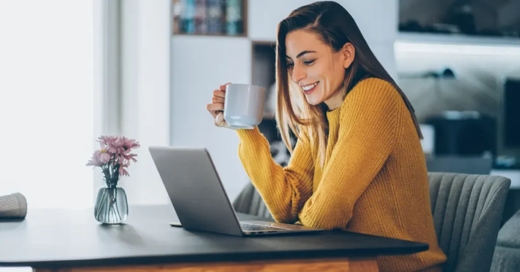 A woman sitting at a table with a laptop and a cup of coffee, smiling, suggesting a comfortable and enjoyable remote working environment.