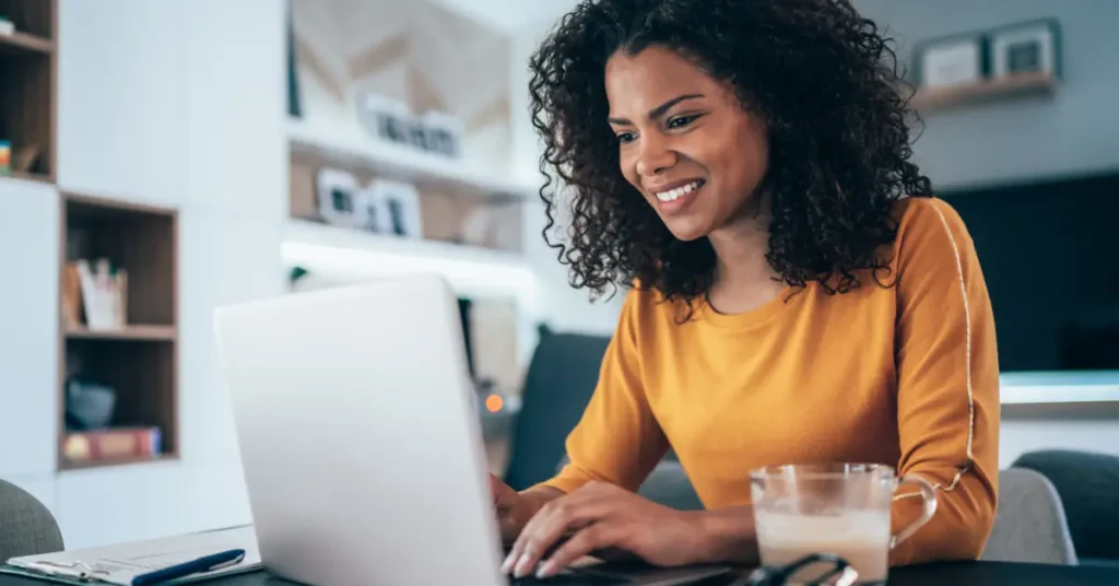 Another smiling woman working on a laptop, wearing a yellow top, indicating a cheerful work-from-home setting. She is looking for remote finance jobs.