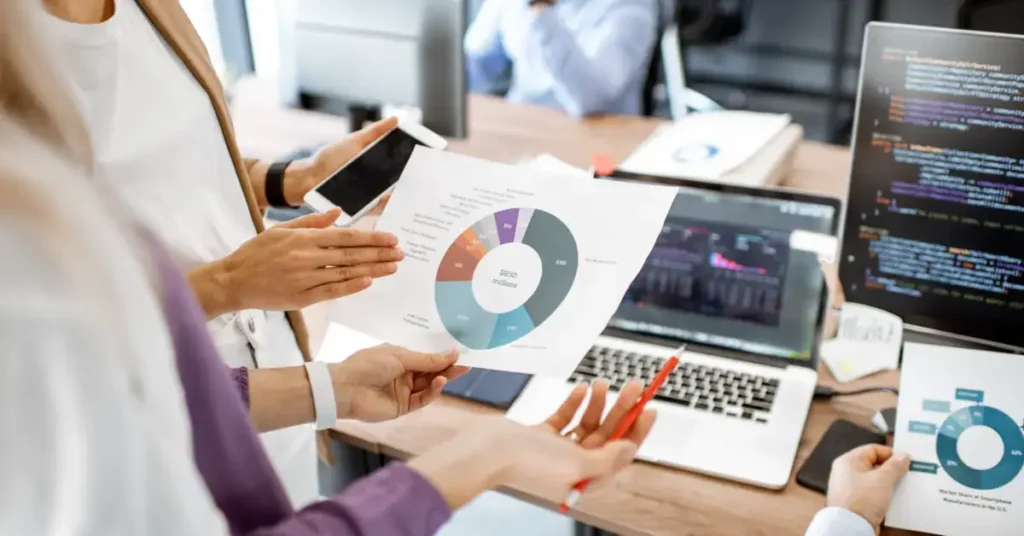 A person holding a tablet and a printed pie chart, with a laptop showing code on the screen in the background. They are used in strategic finance.