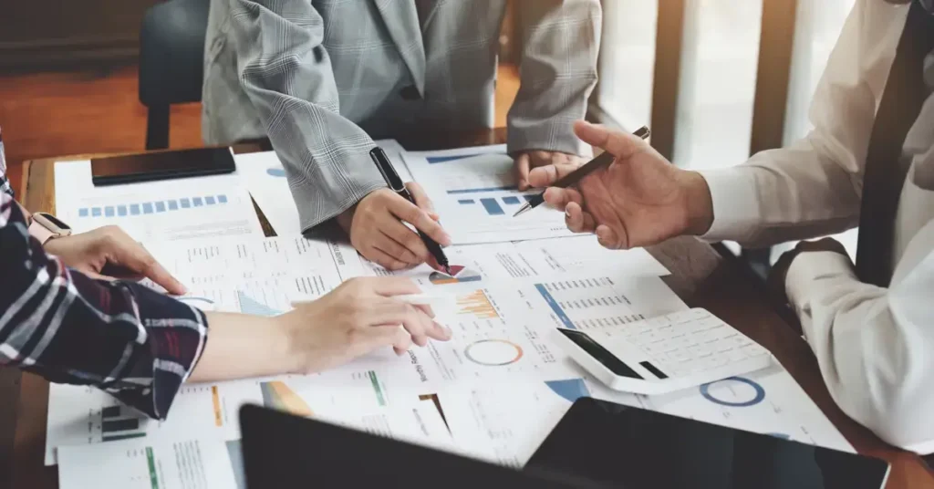 A business meeting with documents and laptops on the table, and a person explaining a point in a strategic finance meeting.