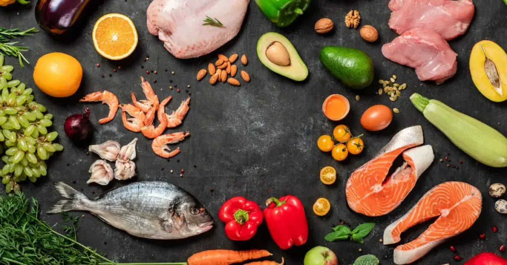 A selection of whole foods spread out on a dark surface, featuring fish, poultry, vegetables, and fruits. Paleo meals contain them