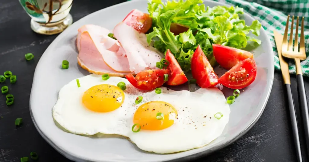 A breakfast plate with two fried eggs, ham, lettuce, and sliced tomatoes. Breakfast idea for budget paleo meals