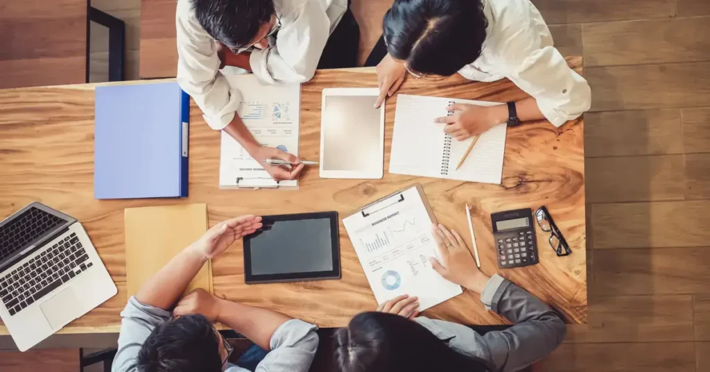 Overhead view of four people around a wooden table with laptops, tablets, documents, and a calculator. humans discussing behavior problems in budgeting