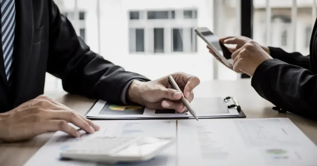 Two individuals at a desk analyzing data on a tablet and documents, with one person pointing at the tablet screen. humans discussing budgeting tablets.